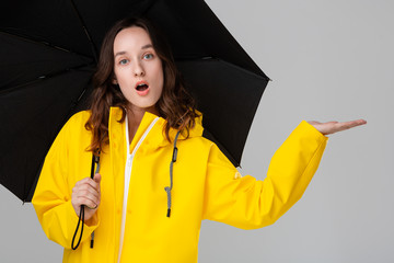 Surprised brunette girl hiding under black umbrella dressed into yellow raincoat. Model checking if rain has started.
