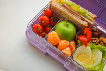 Lilac lunch box with useful food for lunch and snack: sandwich, vegetables, fruits, nuts and eggs on a  white background with space for text. Concept of healthy food, snack for adults and kids