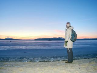 Woman in warm long jacket stay on sandy beach at frozen lake or river. Winter chilly morning with...