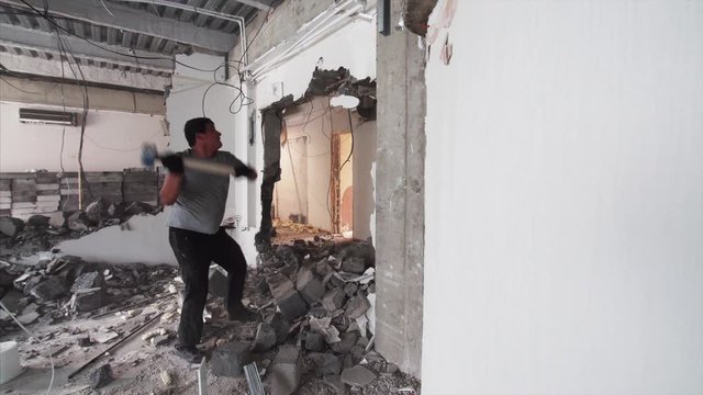 A strong man in gloves is destroying a wall in apartment using heavy hammer. Demolition work in the house. 