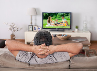 Man sitting on a sofa watching tv with hands folded behind his head