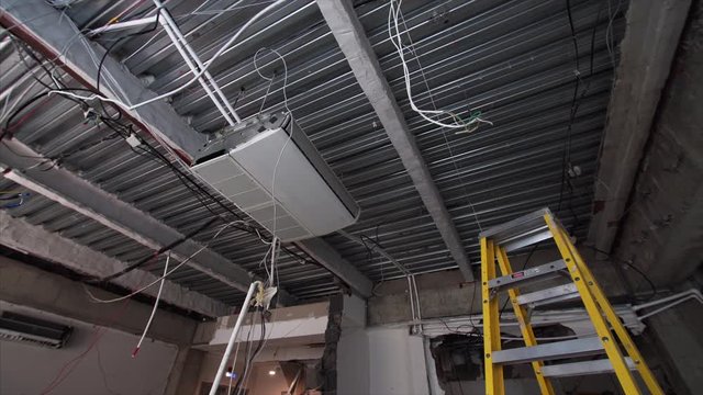 Sliding view of big air conditioner on the ceiling and laddre in the room under construction. Wires are hanging from ceiling