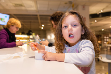 cute little girl is sitting at the table in a restaurant and she is eating an ice cream
