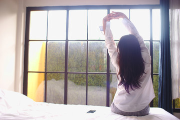 A woman wearing a boyfriend's white shirt is stretching oneself in bed after waking up