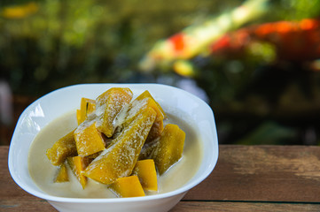 Yummy pumpkin in sweet coconut milk in white bowl, popular Thai dessert on wooden table with blurred fancy carp fish in pond background