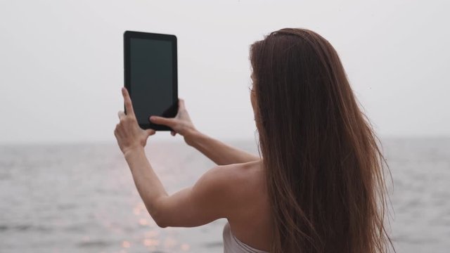 Young woman with long hair in a white dress takes pictures on a tablet