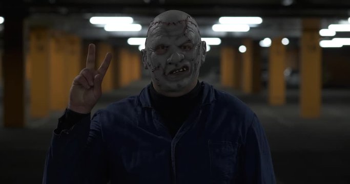 Man in working blue robe and disgusting leather mask stands in front of camera, looks into lens and raises his hand, shows victory sign, scary look, man prepares for Halloween, looks repulsive