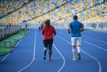 Man and young disabled woman with the prosthetic leg running on the track of the stadium, evening light