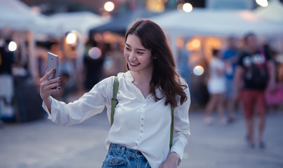 Fototapeta premium Happy traveller young woman using mobile phone to taking selfie at night street market with light bokeh background in Bangkok Thailand.