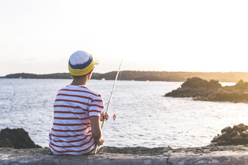 Back view of a young fisherman with with fishing pole in hand looking the sea. Sailor boy with wool cap and a striped t-shirt enjoying a summer evening. Lifestyle, outdoors, fun and carefree concept