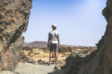 Back view of a teen, standing between lava rocks, looking at the horizon. Trendy boy looks enthusiastically at the view of the Teide National Park. Freedom, travel, tourist and outdoors concept.