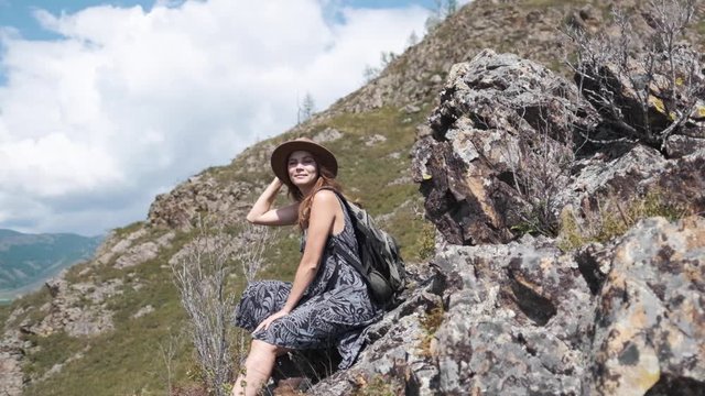 Attractive brunette girl in a hat with a backpack sits on a stone in the mountains looks at the nature