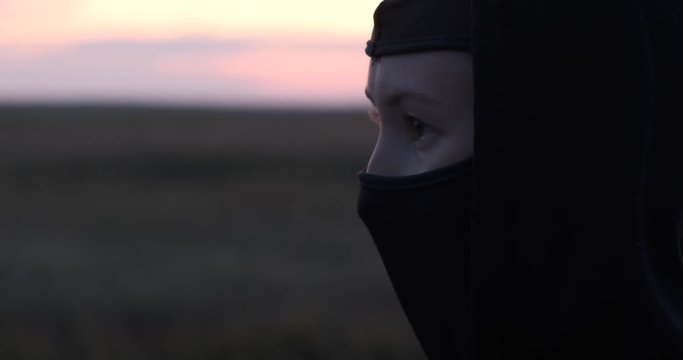 Girl with beautiful eyes looks into space, black balaclava and hood are put on her head, she is standing in open field, sun is setting, evening, inner confidence, readiness to resist, impending unrest