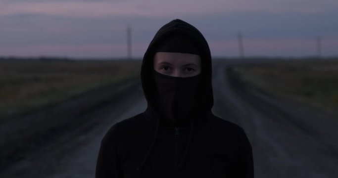 Girl looks into space in front of herself, black balaclava and hood are put on her head, she is standing on country road alone, girl takes off her mask with sharp movement, looks at camera with call