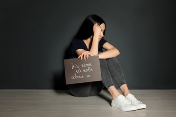 Young woman holding card with words IT'S TIME TO TALK ABOUT IT while sitting near grey wall