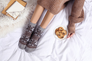 Woman with cookies wearing knitted socks on white fabric, top view. Warm clothes