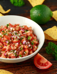 Mexican Tomato Salsa in white bowl with lime, red onion, jalapeno pepper, parsley and tortilla chips on wooden table