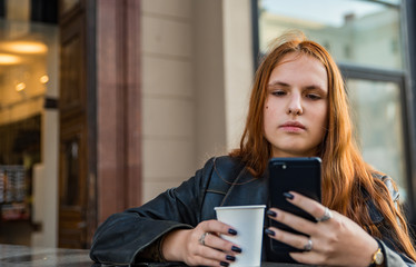 portrait of young teenager redhead girl with long hair with smartphone and cup of hot coffee at city street cafe terrace