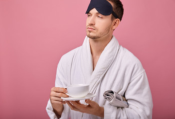 Un sleepy man in a sleeping mask on head in a white coat with a newspaper under his arm, smiling at the camera with a cup of coffee in hands. Awakening, morning mail. Copy space. Looking sideway.