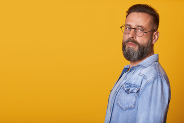 Image of handsome middle aged bearded man with beard, standing and looking directly at camera, posing over yellow background, wearing senim jacket. Copy space for advertismant or promotion text.