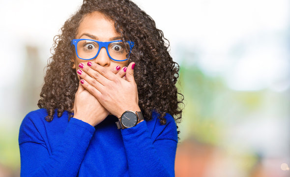 Young beautiful woman with curly hair wearing glasses shocked covering mouth with hands for mistake. Secret concept.