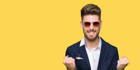 Young handsome elegant man wearing sunglasses and fashion blazer celebrating surprised and amazed for success with arms raised and open eyes. Winner concept.