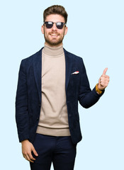 Fototapeta premium Young handsome elegant man wearing sunglasses doing happy thumbs up gesture with hand. Approving expression looking at the camera with showing success.