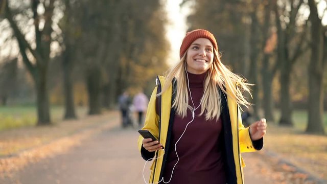 Smiling and happy blonde woman walking in the autumn park. Holding mobile phone, listening to music and enjoying life. Handheld shot. Slowmotion.