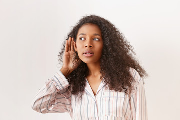 Snoopy nose young dark skinned woman in striped night gown eavesdropping, keeping hand at her ear...