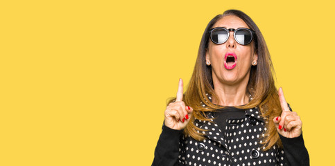 Obraz premium Beautiful middle age woman wearing sunglasses amazed and surprised looking up and pointing with fingers and raised arms.