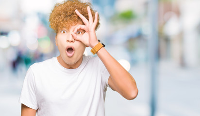 Young handsome man with afro hair wearing casual white t-shirt doing ok gesture shocked with surprised face, eye looking through fingers. Unbelieving expression.