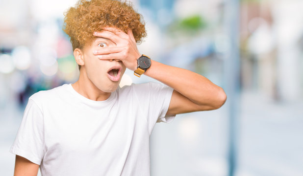Young handsome man with afro hair wearing casual white t-shirt peeking in shock covering face and eyes with hand, looking through fingers with embarrassed expression.