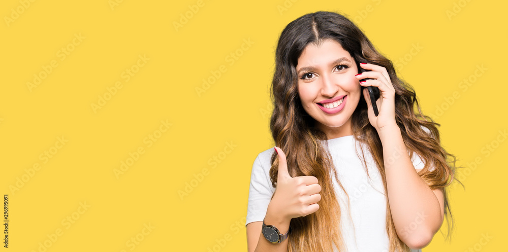 Wall mural Young adult woman taking on the phone happy with big smile doing ok sign, thumb up with fingers, excellent sign