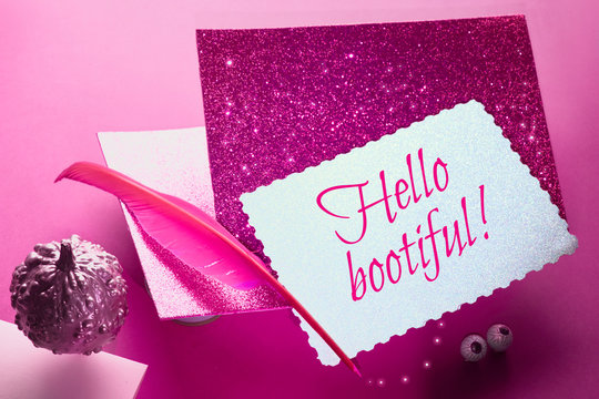 Creative purple and pink Halloween background with levitating pink pin quill, stack of glittering paper and decorative pumpkins painted metallic pink. Text "Hello bootiful"