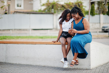 Two african american dark skinned friends female. One of them plus size model, second slim. Having fun and spending time together. Look at mobile phone.