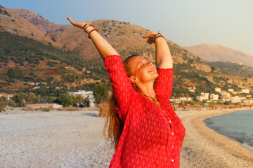 A beautiful young Caucasian woman in a pink blouse with red hair smiles and enjoys the sunset against the background of the Mediterranean Sea and the mountains on the coast of Albania.