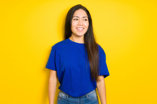Beautiful brunette woman wearing blue t-shirt over yellow isolated background looking away to side with smile on face, natural expression. Laughing confident.