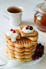 A pile of freshly baked homemade waffles sprinkled with crean and with berries on the top on the table and tea utensils in the background
