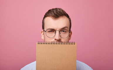 A young student in a gently blue shirt with glasses covered his face with a brown loose-leaf...