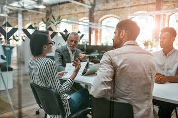 Back view of young business woman holding financial report and discussing something with colleagues while sitting at the office table together
