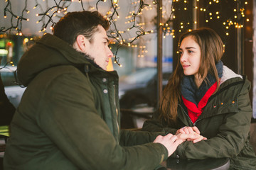 Theme love holiday Valentines Day. pair college students, Caucasian heterosexual lovers in winter, sit table of street cafe against the background window lights. Emotion romance of happiness and love