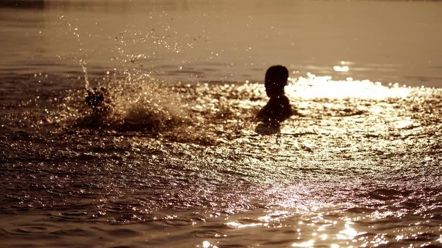 Little kids are playing in water and making splash; Children are swimming in lake or river and having fun with drops; Two brothers rollick at sunset. Monochrome video. Side view.