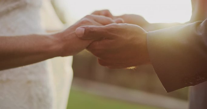 Close up detail shot of diverse bride and groom holding hands for their wedding vows at sunset, bride and groom holding hands getting married with sun flare