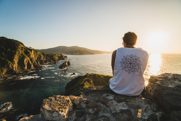 Back of a young traveler sitting on a cliff by the ocean coast watching a beautiful sunset.