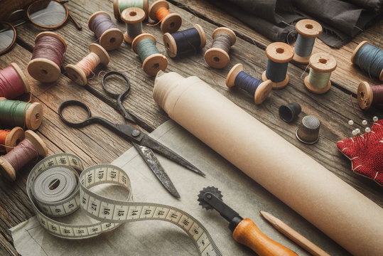 Sewing Items: Retro Tailoring Scissors, Measuring Tape, Thimble, Wooden Spools Of Thread, Patterns On Paper, Cutting Knife, Cushion For Including Pins And Needles. Top View.