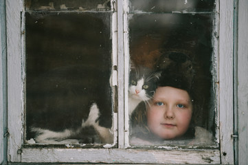 Portrait of little plump girl with her lovely kitten looking out of old window in village house.