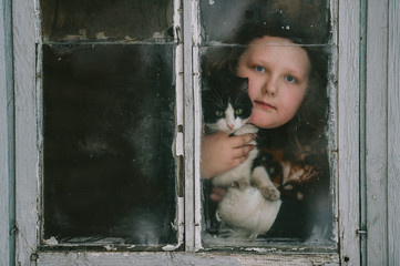 Portrait of little plump girl with her lovely kitten looking out of old window in village house.