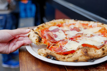 Close up view of hand pick and hold piece of traditional Parma Ham Pizza made with classic wood fired oven, on top with Parmesan cheese, serve on white plate and wooden table on outdoor street market.
