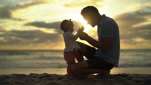 Beautiful baby girl with her father walking on summer beach. Sunrise on the sea bank. Slow motion of playing of small family.