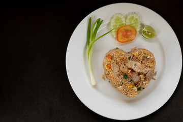 Fried rice with pork a heart shaped dish with vegetable side dishes on a white tile on a black background.